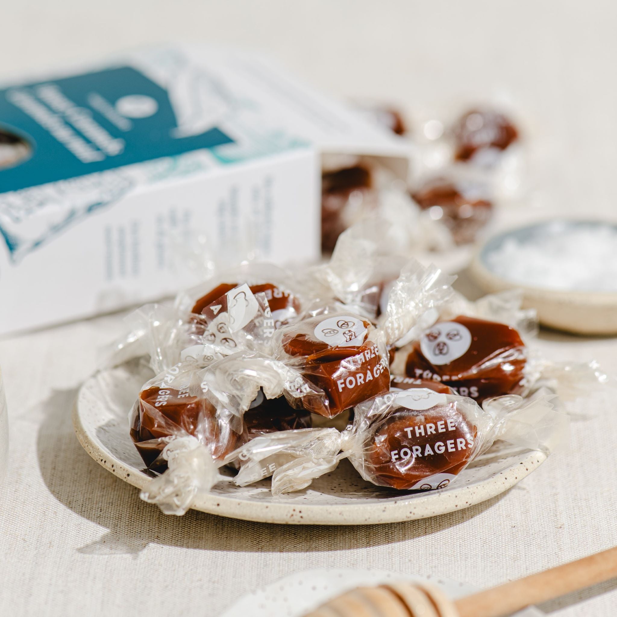A dish of wrapped honey caramels amid a dish holding a honey dipper, a dish with coarse salt, a box of sea salt honey caramels, and a salt well