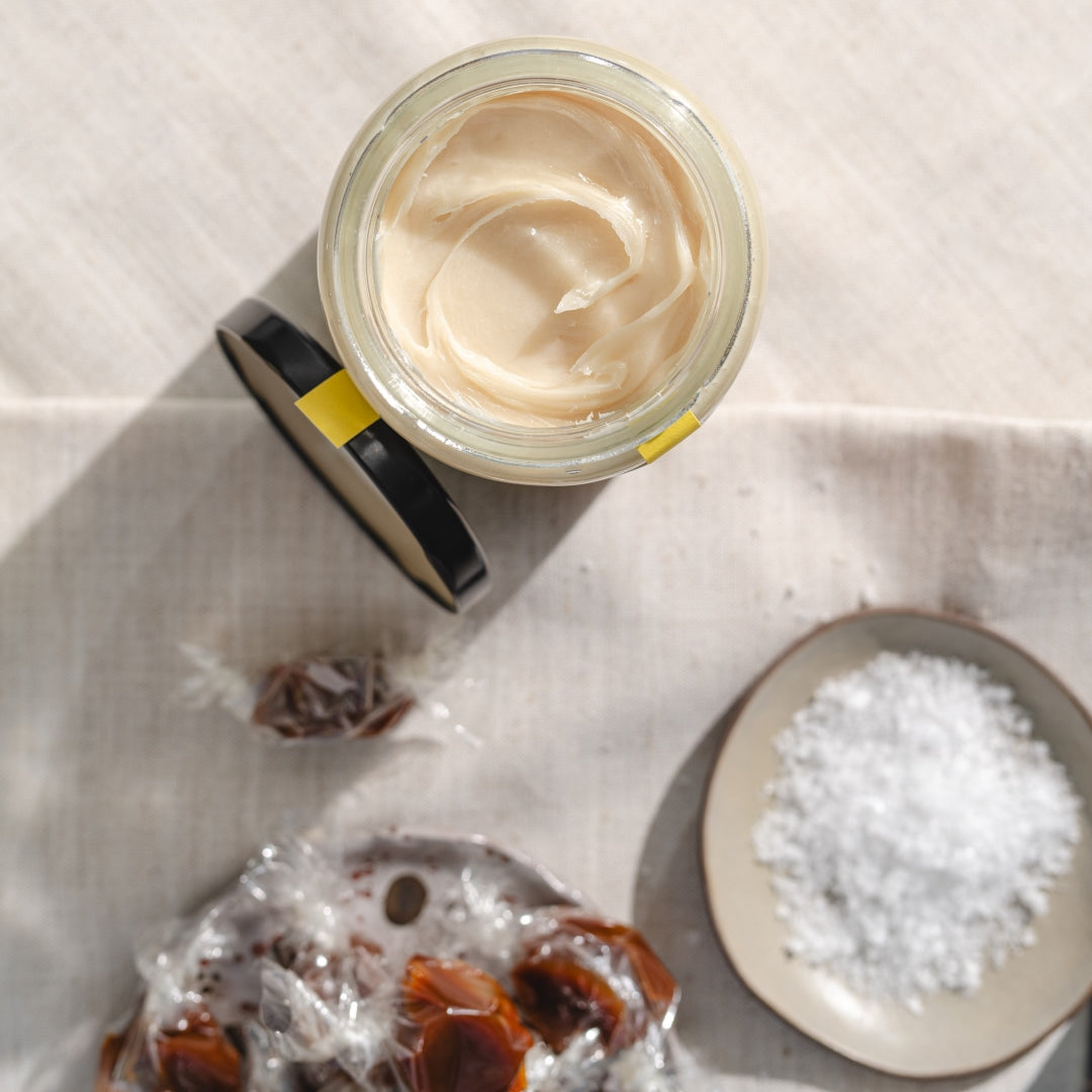 An opened jar of Three Foragers creamed honey next to a dish of coarse salt and a dish of wrapped honey caramels