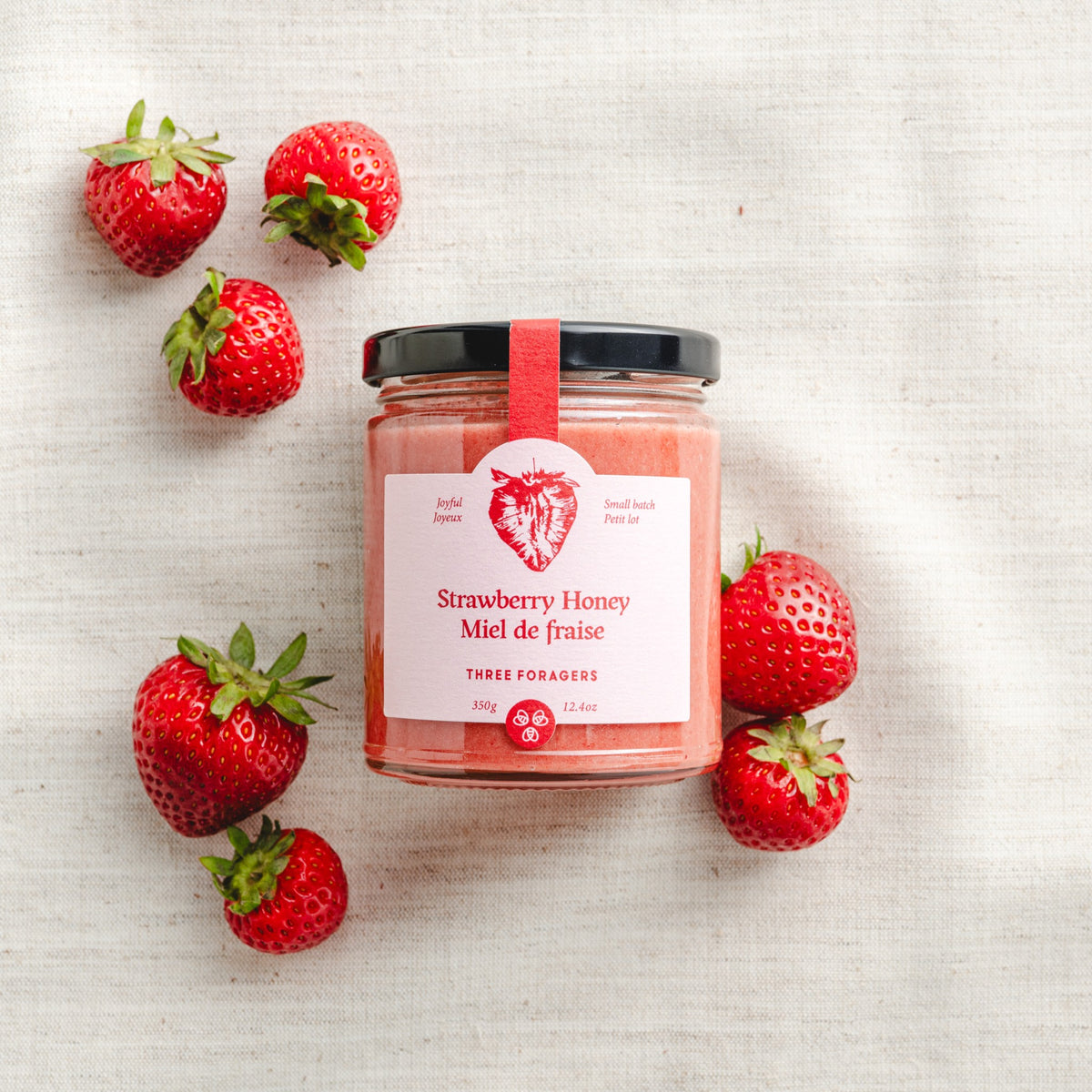 A jar of Three Foragers Strawberry Honey amid some strawberries