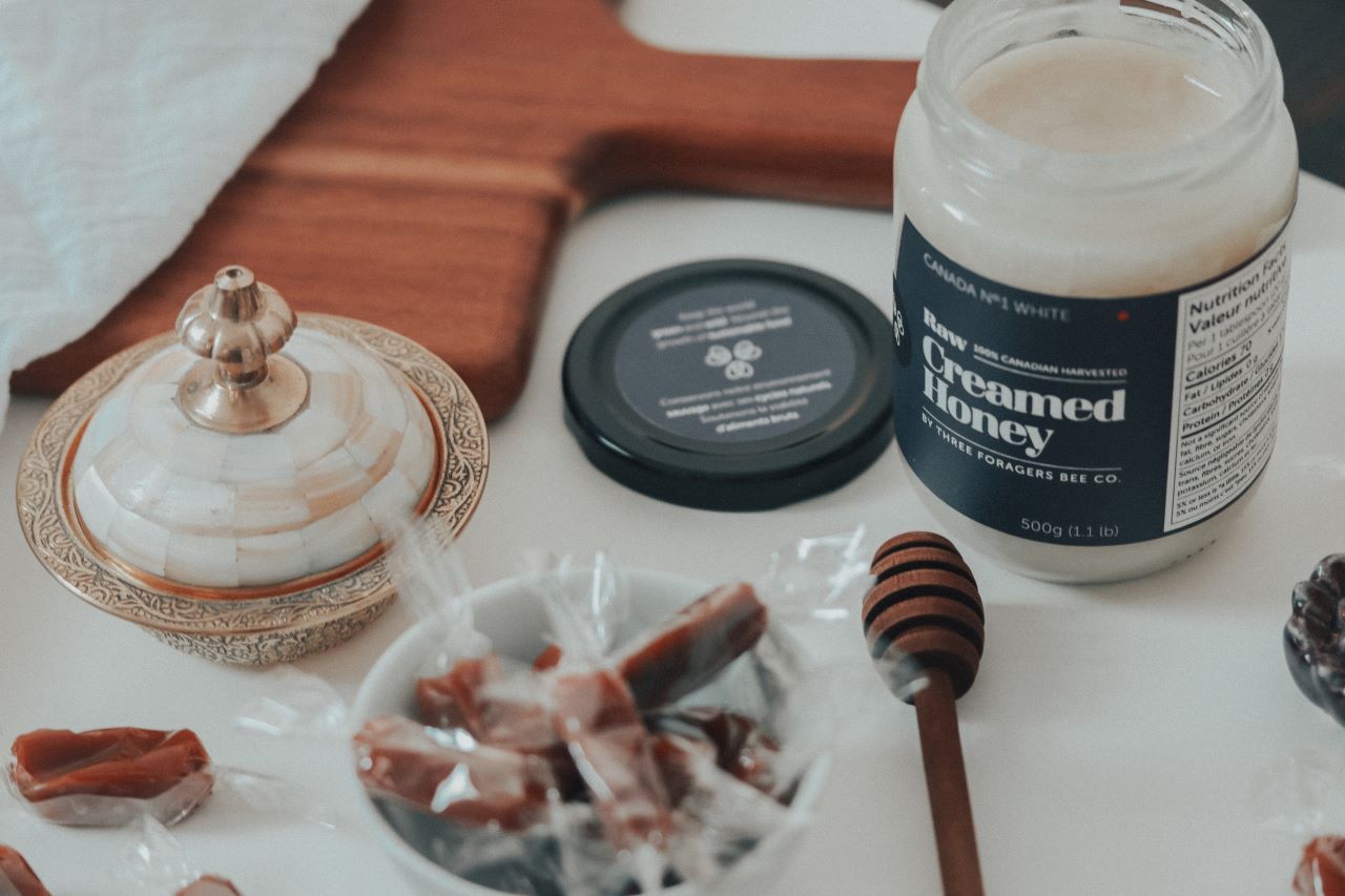 (Left to right) A decorative dish, a dish of wrapped honey caramels, a honey dipper, and a jar of opened Creamed Honey